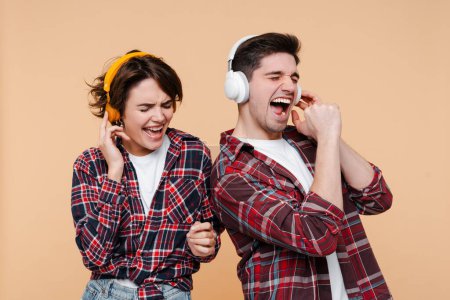 Foto de Emocional pareja de amigos elegantes con auriculares escuchando música, cantando canciones y disfrutando aislados en un fondo beige - Imagen libre de derechos