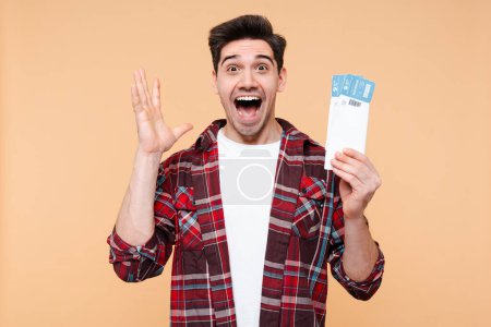 Photo for Excited man, passenger tourist traveler going for weekend getaway, screaming to camera, posing over beige background with a passport and airplane ticket, awaiting to board the flight - Royalty Free Image