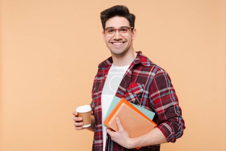 Photo for Portrait of smiling man, student holding coffee and books looking at camera isolated on beige background. Education concept - Royalty Free Image