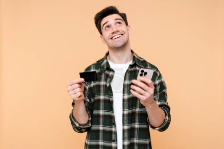 Photo for Portrait dreamy smiling man holding mobile phone, credit card and enjoying shopping online with sales isolated on beige background. Internet store, electronic money concept - Royalty Free Image
