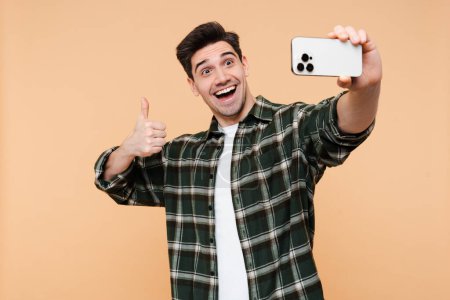Téléchargez les photos : Homme souriant prenant selfie montrant signe cool avec doigt regardant la caméra sur le téléphone mobile, isolé sur fond de couleur beige - en image libre de droit