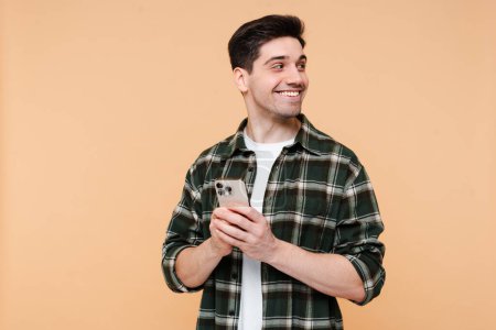 Photo for Portrait of handsome smiling man holding mobile phone shopping online with sale, looking away isolated on beige background. Happy hipster guy using mobile app ordering food, copy space - Royalty Free Image