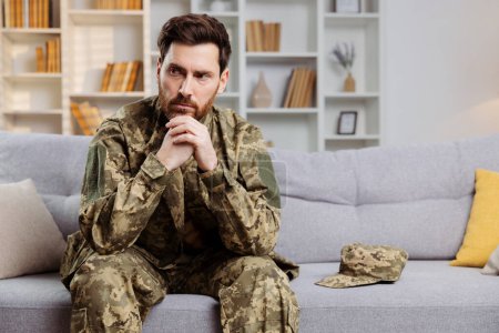 Photo for Soldier returning home theme. Distressed man in Ukrainian army gear seated by himself on a couch. Emphasizes concepts of sorrow, mental strain, and PTSD - Royalty Free Image