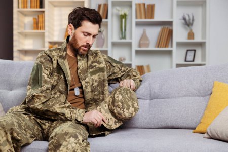Photo for Soldier facing homecoming challenges concept. Distraught man in Ukrainian military gear, isolated on a couch, reflecting on family sorrows and PTSD struggles - Royalty Free Image