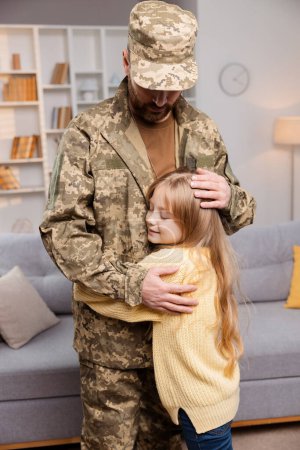 Foto de Sincera escena del regreso a casa del soldado se desarrolla como papá en traje militar envuelve los brazos alrededor de la alegre hija, de pie en el calor de su casa. Soldado aprecia momento de reunirse con sus seres queridos - Imagen libre de derechos
