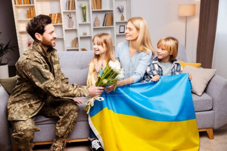Téléchargez les photos : Dans un foyer confortable, le père en équipement militaire ukrainien se réunit avec sa famille sur leur canapé, offrant des fleurs à sa femme, tandis que leurs enfants, tenant le drapeau ukrainien, montrent leur amour et leur excitation vivement - en image libre de droit