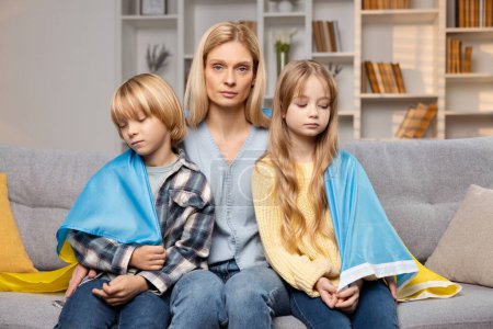 Photo for A Ukrainian mother, sad and protective, holds her children close, enveloped in their country's flag while seated. Their downcast faces reveal a deep longing for home - Royalty Free Image