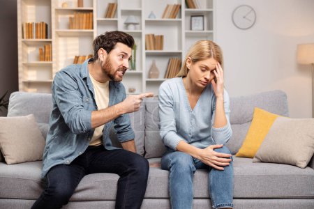 Photo for In their living room, a woman holds her head in despair while her husband yells, pointing fingers, illustrating the desperate need for support in abusive family dynamics - Royalty Free Image
