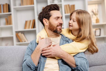 Téléchargez les photos : Scène chaleureuse à la maison, où une fille souriante embrasse son père par derrière alors qu'il se détend sur le canapé confortable, mettant en valeur le lien de la famille dans une atmosphère sereine à la maison - en image libre de droit