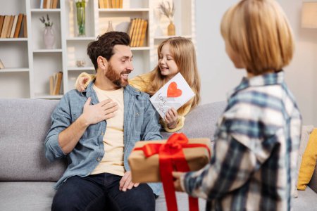Photo for Joyful Father's Day moment captured at home, with excited kids presenting gifts to their astonished dad lounging on living room sofa, showcasing blissful family scene in a perfect domestic setting - Royalty Free Image