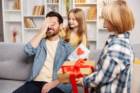 Photo for Celebrating Father's Day, children surprise their dad on the couch with gifts, and the daughter playfully blinds him, engaging in a "guess who" game, filling the living room with laughter - Royalty Free Image