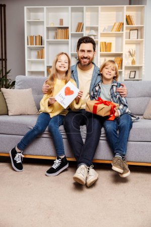Photo for Delighted dad receives gifts from son and daughter on Father's Day, all snuggled together on the living room sofa, sharing a moment of love. All of them looking in camera - Royalty Free Image