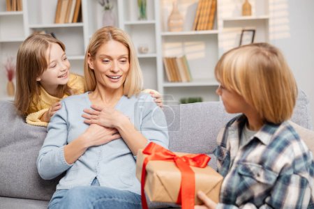 Photo for Warm family moment on Mother's Day with kids giving presents to their astonished mom on the couch, her excitement evident in her wide-eyed, hand-on-chest reaction - Royalty Free Image