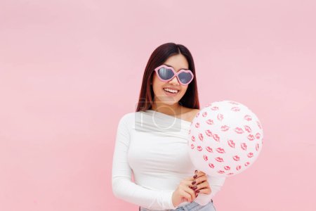 Téléchargez les photos : Heureuse dame asiatique souriante dans des lunettes tenant ballon et regardant la caméra tout en posant isolé sur fond rose. Concept de célébration de la Saint-Valentin - en image libre de droit