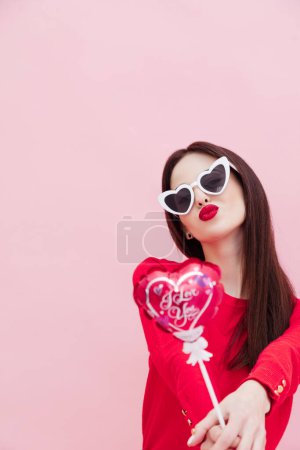 Foto de Vista vertical de la hermosa morena con gafas de sol posando con pequeño globo en forma de corazón aislado sobre fondo rosa - Imagen libre de derechos