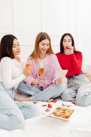 Photo for Cheerful multiracial girls friends sipping alcohol, eating chocolate candies, enjoying celebration holiday sitting at home sofa. Birthday party concept - Royalty Free Image