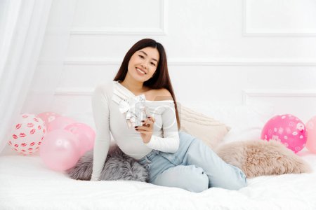 Foto de Linda hermosa mujer asiática sonriente sosteniendo regalo de cumpleaños y sentado en la cama en acogedor apartamento de luz. Concepto de fiestas de celebración - Imagen libre de derechos