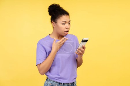 Shocking info concept. Snapshot of a young African American female in a t-shirt, eyes wide at her phone display, isolated on a yellow background