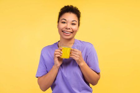 Téléchargez les photos : Concept des émotions humaines. Photo d'une charmante jeune afro-américaine souriante en t-shirt, tenant une tasse de café, isolée sur un fond jaune. Thème énergie du matin - en image libre de droit