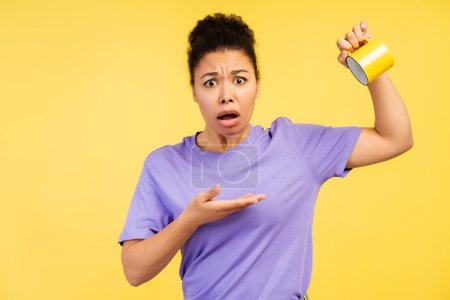 Photo for Emotional portrait concept. Photograph of a young African American woman in a t-shirt, presenting an empty mug with a look of astonishment, inquiring with her gaze, set on a yellow background - Royalty Free Image
