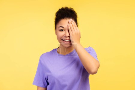 Foto de Concepto de representación de emoción. Captura de una joven y atractiva mujer afroamericana en una camiseta, ocultando juguetonamente un ojo con su mano, sobre un fondo amarillo brillante - Imagen libre de derechos