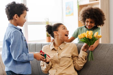 Photo for Delightful Mother's Day moment at home. Snapshot of an ecstatic African American mom on a couch, being presented with unexpected gifts by her little son and daughter - Royalty Free Image