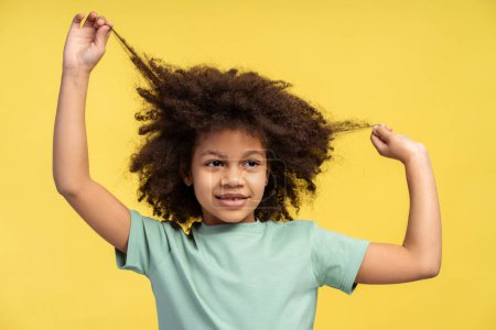 Téléchargez les photos : Heureuse enfant afro-américaine aux cheveux spiralés, vêtue d'une chemise de couleur menthe, lève les bras, tire ses serrures vers le haut, arborant un large sourire, isolée sur un fond jaune vif - en image libre de droit