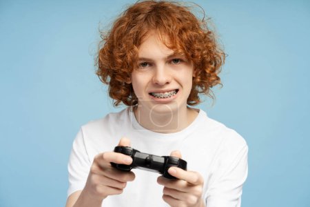 Foto de Muchacho jengibre rizado con bracesplaying videojuego utilizando joystick sobre fondo azul aislado. Mujer muy feliz mirando a la cámara - Imagen libre de derechos
