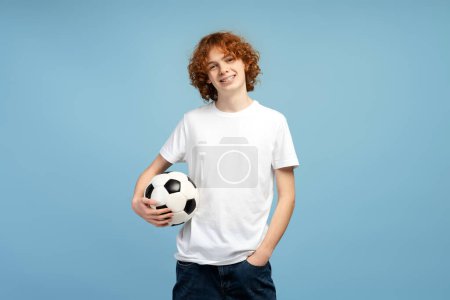 Téléchargez les photos : Portrait d'un adolescent souriant tenant le ballon jouant au football isolé sur fond bleu. Sport, hobby, concept de compétition - en image libre de droit