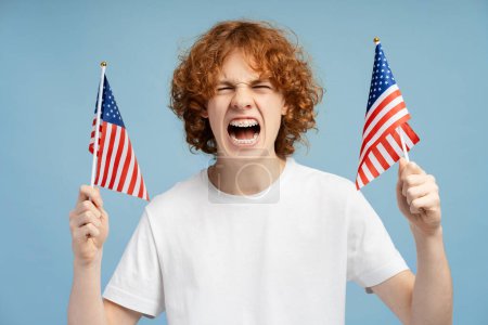 Foto de Jengibre rizado adolescente excitado sosteniendo dos banderas estadounidenses, regocijándose en la victoria aislado sobre fondo azul. Concepto del día de la independencia - Imagen libre de derechos
