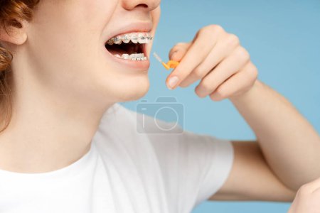 Foto de Primer plano del adolescente jengibre rizado con frenos que sostienen el cepillo de dientes, cepillándose los dientes en el baño en un fondo azul aislado. Concepto de cuidado dental - Imagen libre de derechos