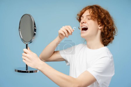 Attractive ginger curly boy looking in mirror, brushing teeth with interdental toothbrush isolated on blue background. Concept of dental care and health 
