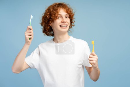 Portrait of handsome curly teenager with braces holding toothbrushes, looking at camera isolated on blue color background. Daily routine and morning hygiene concept 