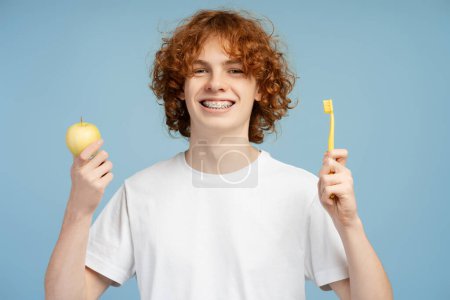 Red haired curly smiling boy with braces in white shirt holding brush and green apple isolated on blue background studio portrait. Healthcare procedures concept