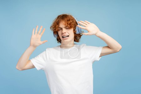 Téléchargez les photos : Portrait d'adolescent attrayant et souriant avec appareil dentaire, écoutant de la musique dans des écouteurs sans fil isolés sur fond bleu - en image libre de droit