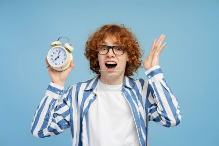 Photo for Shocked emotional boy holding an alarm clock and screaming isolated against blue background. Ginger student late to study - Royalty Free Image