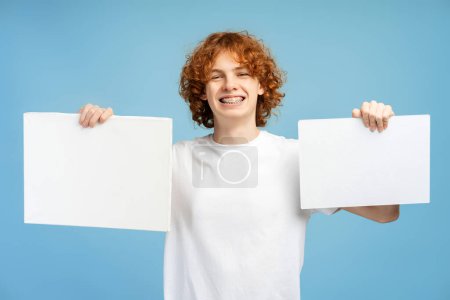 Photo for Happy curly ginger boy wearing white t shirt and with braces holding two empty posters isolated on blue background, mockup. Portrait of smart smiling student with banners - Royalty Free Image