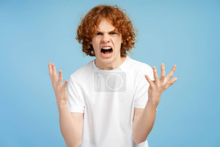 Foto de Chico enojado vistiendo camiseta blanca gritando mirando a la cámara aislada sobre fondo azul. Concepto de publicidad, emociones de la gente - Imagen libre de derechos