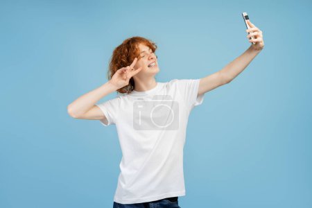 Téléchargez les photos : Garçon gingembre joyeux en utilisant smartphone faire selfie isolé sur fond bleu. Hipster heureux posant caméra de téléphone mobile - en image libre de droit
