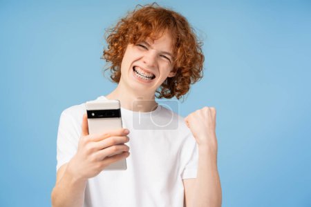 Téléchargez les photos : Adolescent roux heureux tenant mobile hurlant, faisant du shopping en ligne avec des ventes isolées sur fond bleu. Joyeux garçon célébration succès - en image libre de droit