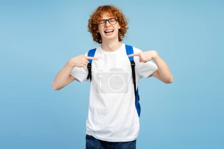 Téléchargez les photos : Un adolescent souriant portant un t-shirt blanc décontracté et un sac à dos se pointant debout avec les yeux fermés isolés sur fond bleu. Maquette - en image libre de droit