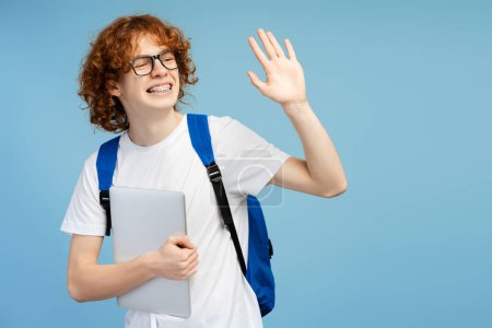 Foto de Atractiva sonriente estudiante rizado de pelo rojo con mochila azul sosteniendo portátil, agitando la mano aislada sobre fondo azul. Concepto educativo - Imagen libre de derechos