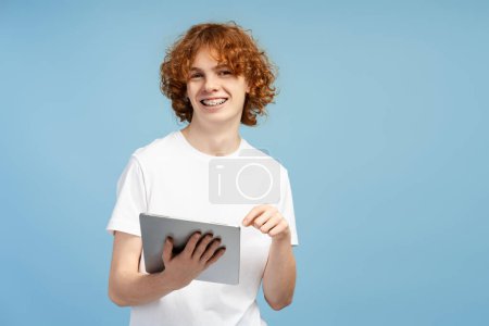 Foto de Sonriente rizado adolescente niño sosteniendo tableta digital aislado sobre fondo azul. Retrato de estudiante feliz inteligente estudiando mirando la cámara, usando tecnología moderna. Concepto educativo - Imagen libre de derechos