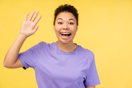Photo for Excited, smiling African American woman waving, greeting, looking at camera isolated on yellow background. Advertisement concept - Royalty Free Image