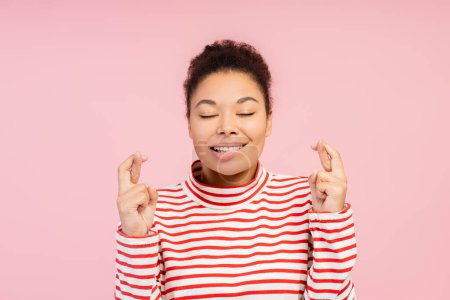 Smiling beautiful African American woman with eyes closed with fingers crossed hoping standing isolated on pink background. Advertisement concept