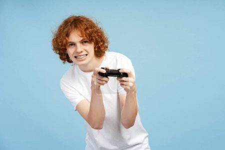 Téléchargez les photos : Un jeune homme concentré aux cheveux roux bouclés et aux bretelles s'engage avec une manette de jeu, isolé sur un fond de studio bleu - en image libre de droit