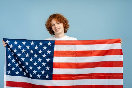 Foto de Sonriente, jengibre rizado adolescente macho con frenos dentales, sosteniendo una gran bandera de EE.UU., colocada sobre un fondo azul. Destaca el orgullo patriótico y el concepto electoral - Imagen libre de derechos