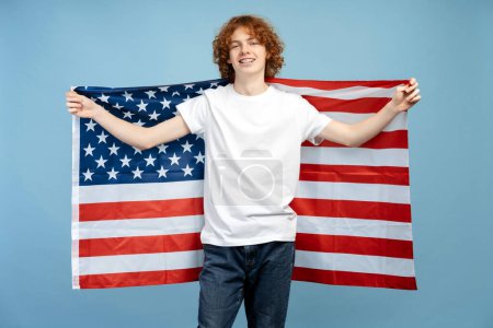 Téléchargez les photos : Photo d'un adolescent roux aux cheveux bouclés et heureux portant des bretelles, en vêtements décontractés, tenant le drapeau des États-Unis derrière son dos, isolé sur un fond bleu. Souligne un concept de visa étudiant - en image libre de droit