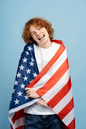 Téléchargez les photos : Joyeux adolescent roux avec bretelles, enveloppé dans un grand drapeau américain, isolé sur un fond bleu. Représente le concept de migration étudiante - en image libre de droit