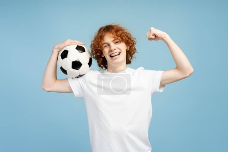 Téléchargez les photos : Photo d'un adolescent roux aux cheveux bouclés et souriant avec bretelles, montrant ses muscles en vêtements de sport, serrant un ballon de football, isolé sur un fond bleu. Concept sportif - en image libre de droit